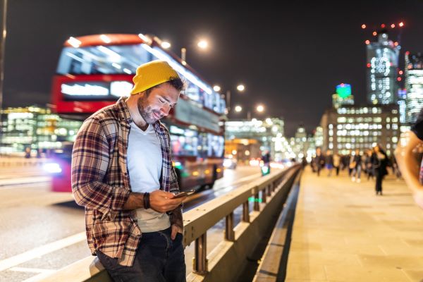 Hombre joven revisando su teléfono en una calle de Londres, leyendo sobre el costo de vida en Inglaterra